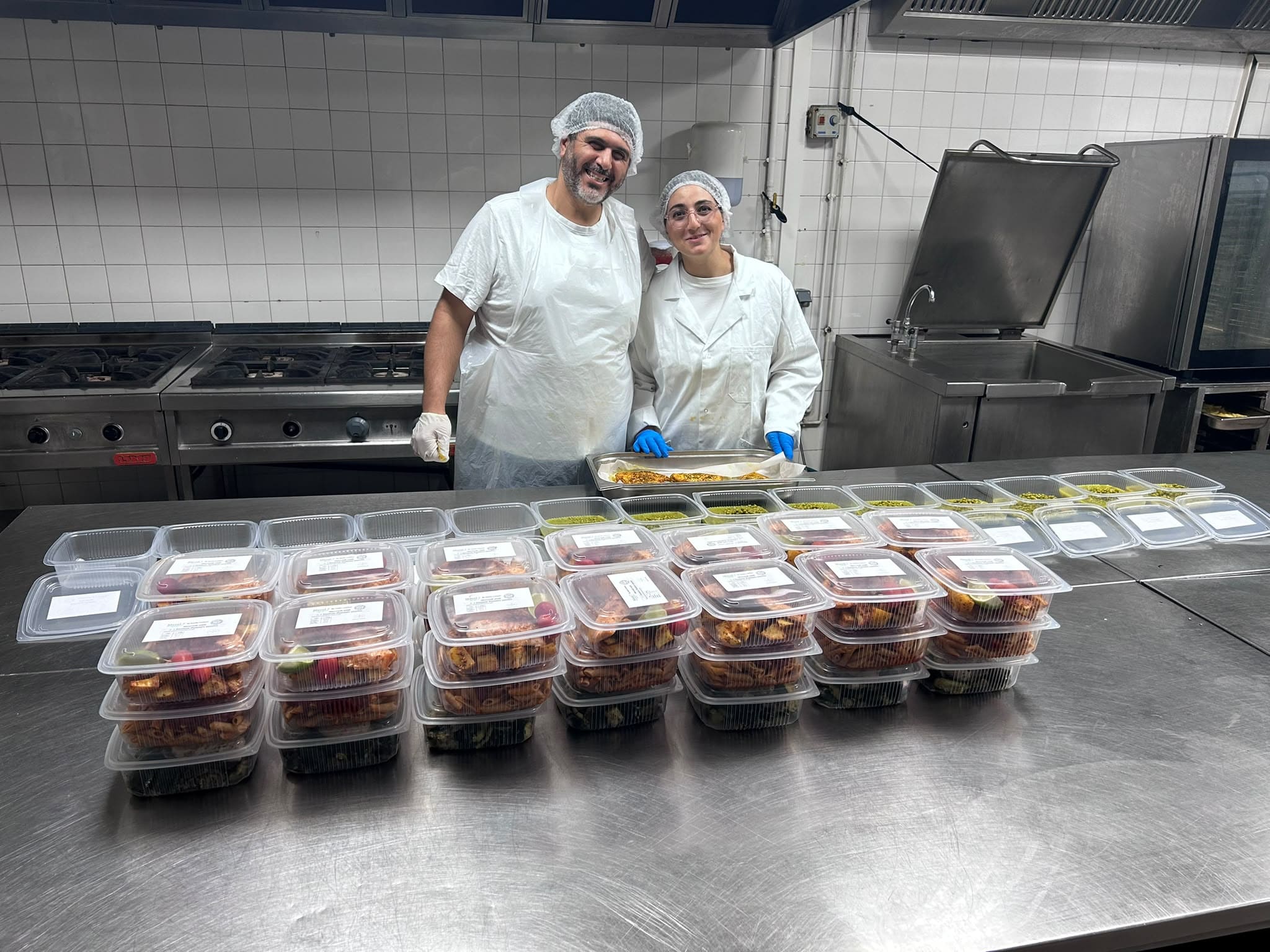 Cuisinier et diététicienne en tenue, devant des barquettes étiquetées alignées sur le plan de travail en inox